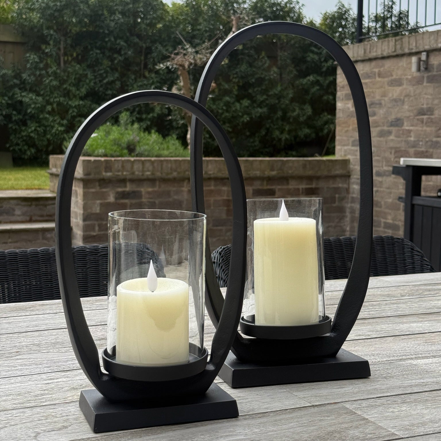Two black metal candle holders with white candles on a wooden table outdoors.