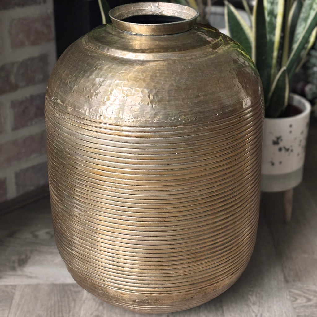 Large metallic vase on a floor with a plant in the background