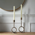 Two candle holders with candles on a wooden surface against a wall with wainscoting.