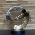 Abstract metal sculpture on a black base against a wooden paneled wall.