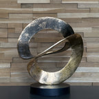 Abstract metal sculpture on a black base against a wooden paneled wall.