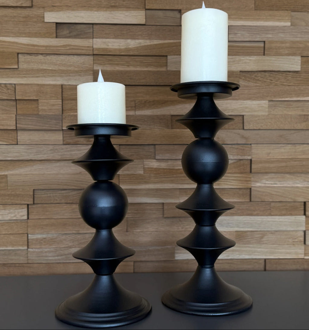 Two black candle holders with white candles against a wooden paneled wall.